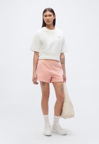 T-shirt blanc en coton à manches courtes avec un logo vert, accompagné d'un short en coton rose clair. Le modèle porte des baskets et des chaussettes blanches, tenant un tote bag crème.
