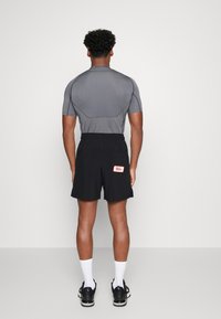 T-shirt de sport gris à manches courtes associé à un short noir avec un écusson logo rouge, porté avec des chaussettes blanches et des chaussures noires.