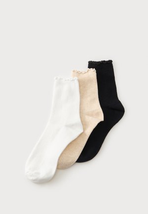 Trois chaussettes courtes côtelées en blanc, beige et noir avec des bords festonnés, posées à plat sur un fond clair.