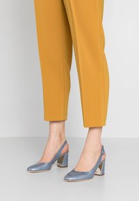 Vaaleansiniset nilkkaketju-korkokengät, joissa on terävä kärki ja paksu metallinen korko, yhdistettynä sinappikeltaisiin nilkkapituisiin housuihin. Sujuva pinta.