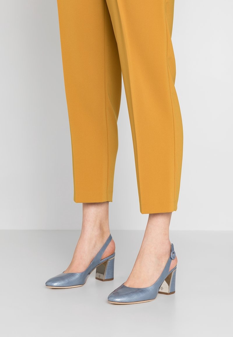 Vaaleansiniset nilkkaketju-korkokengät, joissa on terävä kärki ja paksu metallinen korko, yhdistettynä sinappikeltaisiin nilkkapituisiin housuihin. Sujuva pinta.