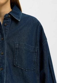Dunkelblaues Denimhemd mit einer Brusttasche, Knopfverschluss und spitzem Kragen. Es hat einen lässigen Schnitt und sichtbare Nahtdetails.