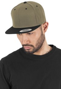 Cappellino snapback verde oliva con visiera piatta nera, texture in tessuto e occhielli neri. Indossato con una t-shirt nera a girocollo.