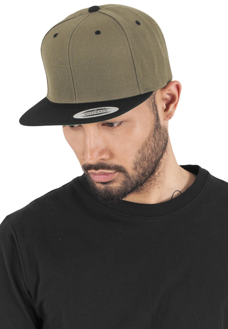 Cappellino snapback verde oliva con visiera piatta nera, texture in tessuto e occhielli neri. Indossato con una t-shirt nera a girocollo.