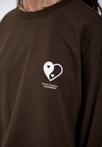 Braunes Baumwoll-T-Shirt mit rundem Ausschnitt, das ein weißes Herz-Logo und den Markennamen auf der Brust aufgedruckt hat. Lockerer Schnitt und glatte Textur.