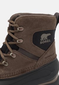 Botas marrones impermeables con parte superior de ante y tejido, acento de malla negra, ojales de metal y diseño de cordones. Logo de Sorel impreso en el lado.