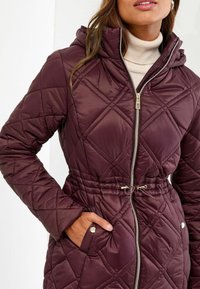 Vrouw in een gevoerde bordeauxrode jas met rits en een koord in de taille, met de hand in de zak, en een beige coltrui eronder.