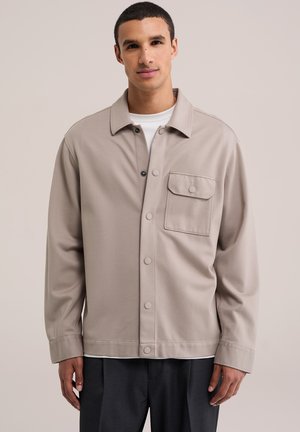 Jeune homme portant une veste-chemise beige à boutons avec une poche poitrine et un pantalon noir, debout devant un fond clair uni.