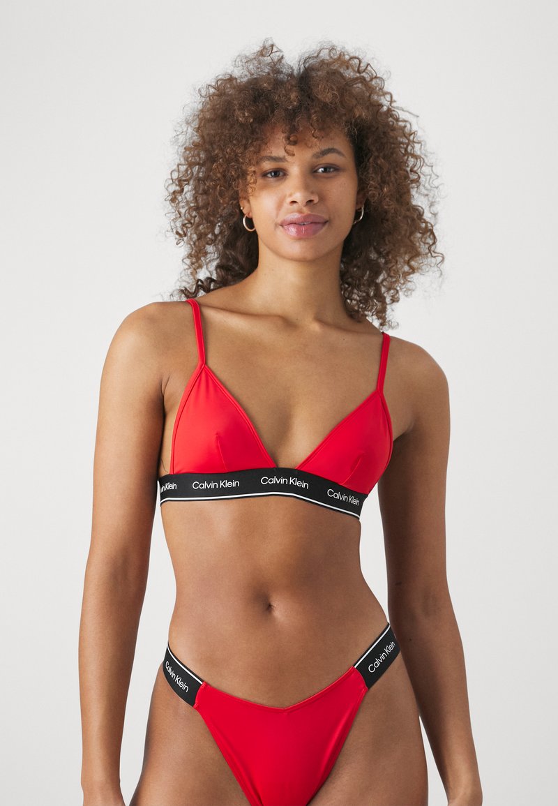 Rød bikini-sett som har en trekantet bh med tynne stropper og et svart elastisk bånd merket "Calvin Klein", supplert med matchende lavt midje truse.