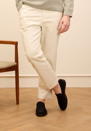 Cremfarbene maßgeschneiderte Hose, entspannte Passform, kombiniert mit schwarzen Wildleder Slippern. Helle Holzböden und ein Holstuhl im Hintergrund.