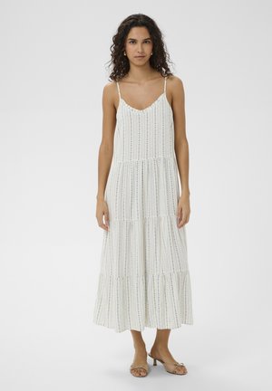 Žena s kučeravými tmavými vlasmi v bielych midi šatách bez rukávov s tenkými modrými vertikálnymi pruhmi a béžovými sandálmi na podpätku, stojaca na bielom pozadí.