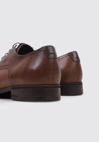 Zapatos de vestir de cuero marrón con una textura suave, forma redondeada y una suela de goma negra. Presentan costuras sutiles y un diseño minimalista.