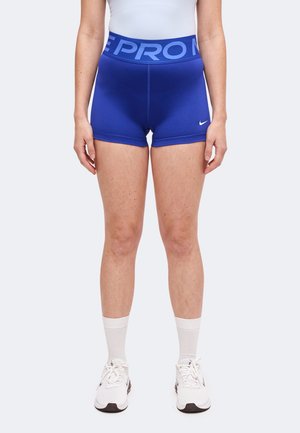 Femme portant un short de sport Nike Pro bleu, des chaussettes blanches, des baskets blanches et un haut blanc, debout devant un fond uni.