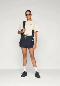 Beige t-shirt med logo, navy shorts med for pockets, grøn snor, hvide sokker og sorte sneakers. Accessoriseret med solbriller og en rygsæk.