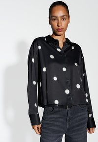 Black, long-sleeve button-up shirt with large white polka dots, a relaxed fit, and glossy texture; paired with dark jeans.