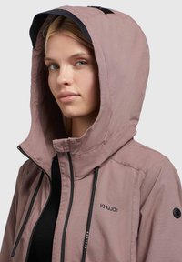 Hellrosa Kapuzenjacke mit schwarzen Akzenten. Verfügt über einen Reißverschluss, verstellbare Kordeln und einen weichen, strukturierten Stoff. Logo-Details sind sichtbar.