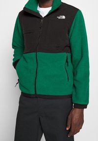 Fleece bunda se zeleným tělem a černými rameny, s předním zipem, dvěma bočními kapsami a kapsou na prsou. Logo na levé straně.