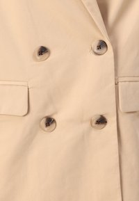 Manteau beige croisé à double boutonnage avec quatre boutons marbrés et poches à rabat de chaque côté.