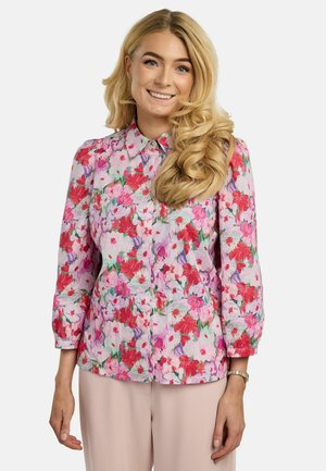 Uśmiechnięta kobieta z długimi, blond lokami, ubrana w różową, kwiecistą bluzkę na guziki i jasne spodnie, na tle jednolitego tła.