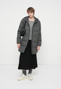 Grå pufferjakke med horisontalt quiltet design, hætte, lynlås foran, parret med en grå sweater og sort plisseret nederdel. Modellen har hvide sneakers på.