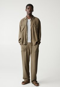 Cămașă maro, texturată, cu două buzunare frontale, tricou gri cu guler rotund dedesubt, pantaloni maro cu șnur asortați și sandale maro.
