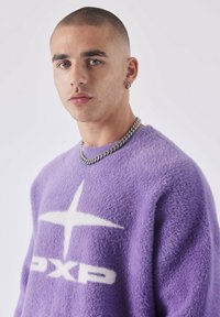 Suéter morado fuzzy con un logo de estrella blanca y el texto "PXP". La tela tiene una apariencia suave y texturizada. Collar de cadena delgada de plata.
