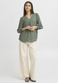 Blusa verde oliva con scollatura a V, tessuto testurizzato e maniche a sbuffo abbinata a pantaloni beige a gamba larga e sandali neri con cinturini.