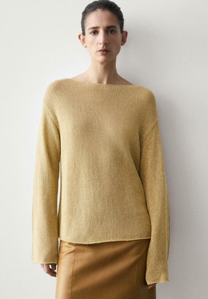 Donna che indossa un maglione beige lavorato a maglia largo con maniche lunghe e una gonna di pelle marrone, in piedi contro uno sfondo chiaro e semplice.