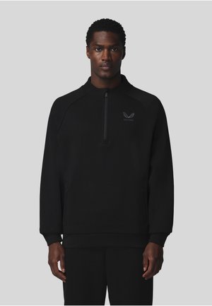 Homme portant un pull noir à manches longues avec une fermeture éclair quatre quarts et un petit logo sur le côté gauche de la poitrine, debout devant un fond gris uni.