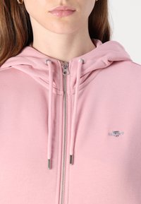Junge Person trägt einen hellrosa Kapuzenpullover mit Reißverschluss, Kordeln, silbernem Reißverschluss und einem kleinen bestickten Logo auf der Brust.
