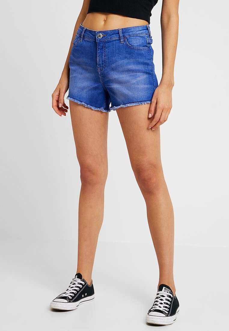 Mujer vestida con pantalones cortos de mezclilla azul con dobladillos deshilachados, top corto negro y zapatillas bajas negras con cordones blancos, de pie sobre un suelo blanco.