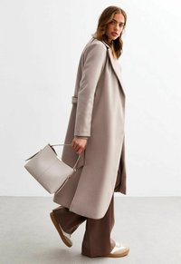 Manteau beige surdimensionné avec un design superposé, associé à un pantalon large marron et un sac gris clair. Présente un tissu lisse et une silhouette structurée.