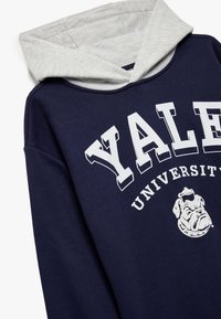 Sudadera azul marino con capucha gris, que presenta un texto blanco que dice "YALE UNIVERSITY" y un gráfico de un bulldog en la parte delantera. Textura suave.