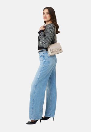 Femme portant une veste à motifs noirs et blancs, un jean large bleu clair, des talons noirs et un sac à bandoulière beige.