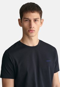 Černé tričko s krátkým rukávem vyrobené z bavlny, s kulatým výstřihem a malým modrým logem GANT na levé hrudi. Jednoduchý a elegantní design.