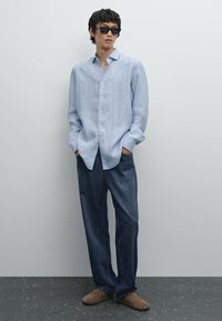 Camicia a maniche lunghe a righe blu chiaro con colletto, abbinata a jeans rilassati blu scuro e scarpe slip-on marroni, indossata con occhiali da sole.