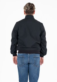 Man met kort haar, gekleed in een zwarte jas en blauwe jeans, staande met zijn rug naar de camera tegen een eenvoudig wit achtergron.