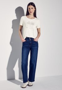 Bílé tričko s krátkým rukávem a metalickým potiskem, v kombinaci s tmavě modrými rovnými džíny. Model má na sobě béžové tenisky. Jednoduché pozadí.