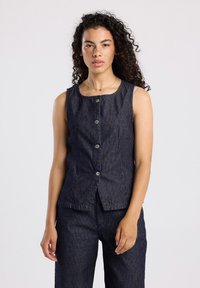 Nuori nainen, jolla on kiharaiset hiukset, pukeutuneena olkaimettomaan, napitettyyn tummansiniseen raidalliseen liiviin ja siihen mätsääviin housuihin, vaaleaa taustaa vasten.