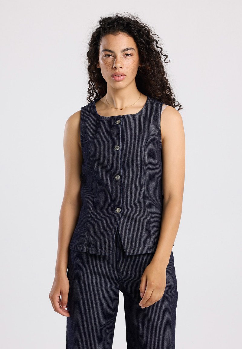 Nuori nainen, jolla on kiharaiset hiukset, pukeutuneena olkaimettomaan, napitettyyn tummansiniseen raidalliseen liiviin ja siihen mätsääviin housuihin, vaaleaa taustaa vasten.