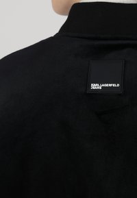 Musta bomber-takki, jossa on teksturoitu kangas, ribbikaulus ja nelikulmainen etiketti, jossa on valkoisella kirjoitettu "KARL LAGERFELD JEANS".