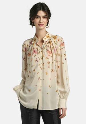 Mujer con blusa larga de manga larga color crema con estampado floral, cuello con volantes, botones al frente, y pantalones negros, de pie frente a un fondo sencillo.