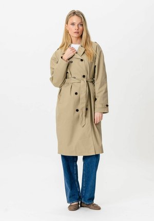 Lyshåret kvinde iført beige dobbeltradet trenchcoat med bælte, blå jeans og brune flade sko stående foran en ensfarvet baggrund.