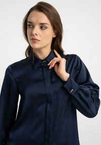 Granatowa satynowa bluzka z kołnierzykiem, długimi rękawami i pojedynczymi mankietami z ozdobami w kolorze złotym. Gładka faktura, elegancki design.