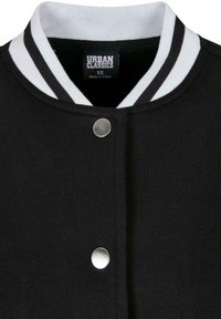 Musta varsity-takki, jossa on valkoraidallinen resoripinta kauluksessa; varustettu hopeisella nepparilla ja pehmeällä kangaspinnalla. Merkissä lukee "Urban Classics, XS."