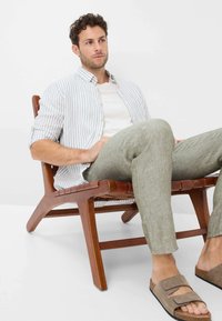 Striped, button-up shirt in white and light blue, with short sleeves. Light beige linen pants and brown slide sandals. Wooden lounge chair.