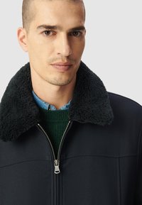 Chaqueta azul marino con cremallera y un suave cuello negro, combinada con un suéter verde y una camiseta de color azul claro. Tela suave y detalle de cremallera visible.