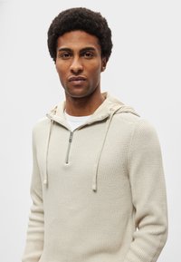 Hoodie bege em malha com fecho de correr no pescoço, apresentando um padrão texturizado e cordões no capuz. Forro interior suave, design casual.