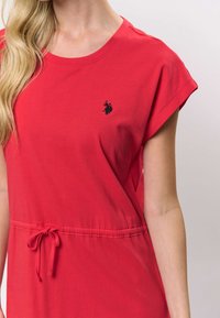 Rochie roșie din bumbac, cu decolteu rotund, mâneci scurte și talie ajustabilă cu șnur. Are un mic logo negru pe piept.