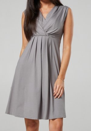 Femme portant une robe grise sans manches, longueur genou, avec un col en V et une taille plissée, se tenant devant un fond uni.